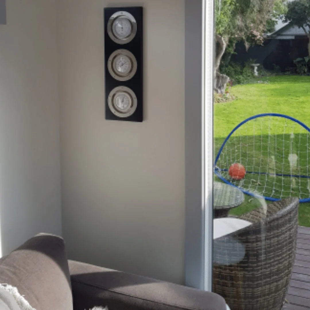 Wall-mounted weather station with barometer, hygrometer, and clock in a living room by a window.