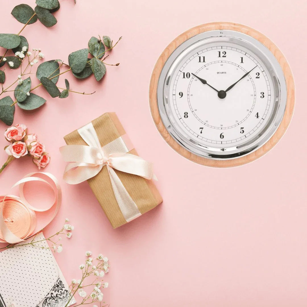 Wall clock with silver trim and wooden frame on pink background, next to gift and flowers