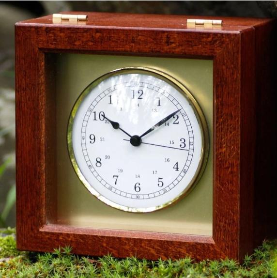 Nautical clock with white dial in a square wooden case, displayed outdoors on moss.