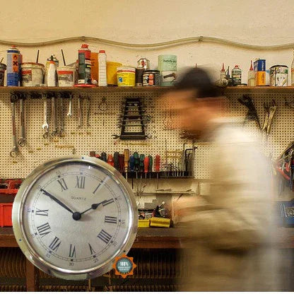 Large wall clock with Roman numerals in a workshop with tools and blurred person walking by