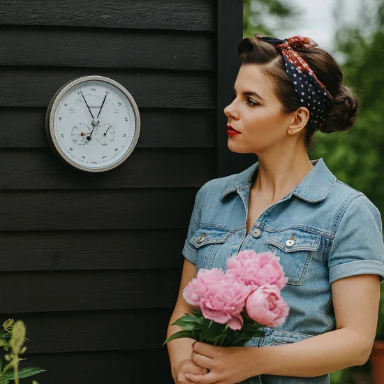 Woman with pink flowers standing beside outdoor wall-mounted weather barometer and hygrometer.